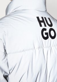 Veste matelassée en matériau gris clair avec un col montant et un logo "HUGO" noir au dos. Texture lisse avec un design matelassé.