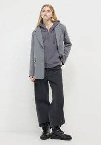 Blazer grigio sovrapposto a un hoodie grigio con zip, abbinato a pantaloni cargo neri larghi e stivali neri con lacci. Trame lisce e strutturate.