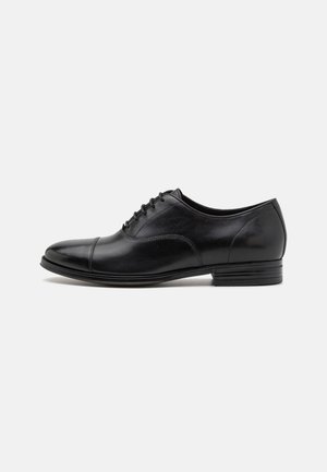 Zapato oxford de cuero negro con un acabado suave, punta redonda, cierre con cordones y tacón bajo; presenta acentos de costura en contraste.