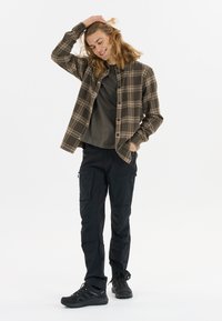 Geruite button-up shirt in bruin en beige, gelaagd over een donkergroen t-shirt; gecombineerd met zwarte cargo-broeken en zwarte wandelschoenen.