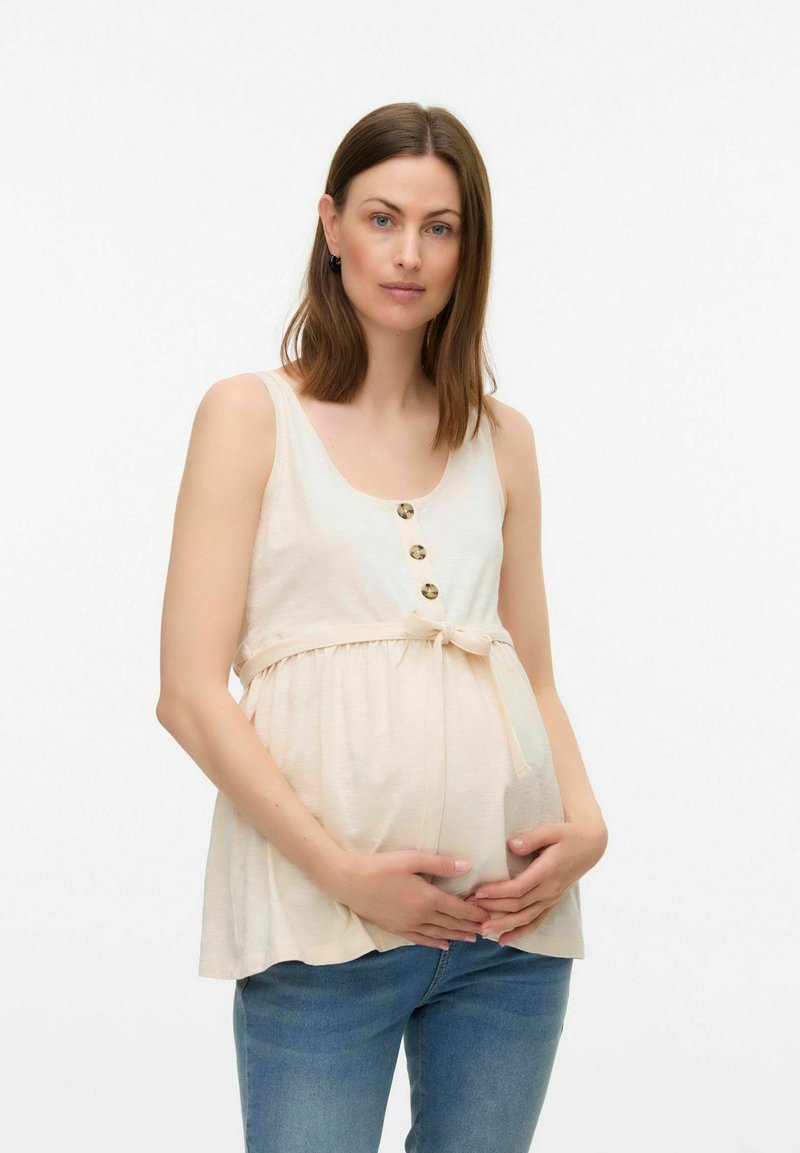 Ljusbeige graviditetstopp med rund halsringning, knappar och en rosettdetalj vid midjan, tillsammans med blå denimjeans.