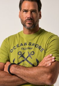 Zelené bavlněné tričko s výrazným námořnickým grafickým designem, který zahrnuje text "OCEAN SHORE OLD SHIP" a motivy kotvy. Krátké rukávy, kulatý výstřih.