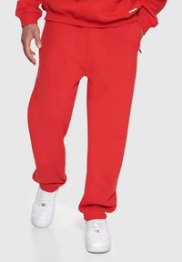 Røde sweatpants lavet af blødt stof, med en afslappet pasform, manchetter ved anklerne og sidelommer, parret med hvide sneakers.