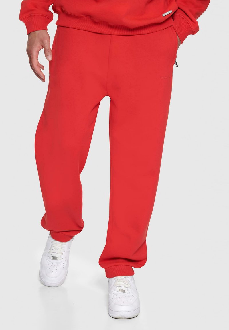 Røde sweatpants lavet af blødt stof, med en afslappet pasform, manchetter ved anklerne og sidelommer, parret med hvide sneakers.