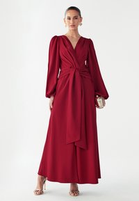 Pikk punane kleit mähisdisainiga, millel on punnitatud pikad varrukad, vöökohal seotav detail ja sile kangapind. Kaunistatud kuldse tooniga riistvaraga.