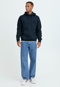 Felpa con cappuccio blu navy con tasca frontale e coulisse, abbinata a jeans azzurri. Tessuto strutturato con logo circolare sul petto. Scarpe da ginnastica bianche.