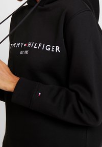 Czarna bluza z kapturem wykonana z mieszanki bawełny, z kieszenią typu kangurka. Posiada białą haftowaną aplikację logo "TOMMY HILFIGER" oraz mały akcent logo na rękawie.