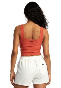 Kobieta z długimi kręconymi włosami, ubrana w pomarańczowy top bez rękawów i białe szorty z prążkowanego materiału z tylną kieszenią, stojąca tyłem na białym tle.