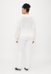 Weißer gestrickter Pullover mit lockerer Passform und gerippten Bündchen, kombiniert mit weißen, gerade geschnittenen Hosen und schwarzen, mit Nieten verzierten High Heels.