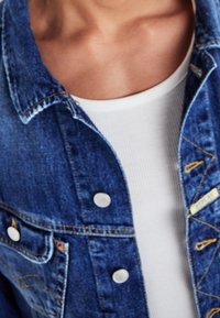 Person wearing a blue denim jacket over a white ribbed tank top, showing the collarbone and upper chest area.