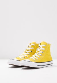 Gule sneakers med høj top lavet af lærred, med hvide snørebånd, gummisåler og kontrasterende sorte striber langs sålen.
