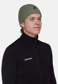 Green ribbed knit beanie with a logo patch, worn by a person in a black fleece jacket featuring a zip and white branding.