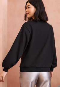 Crna dukserica s opuštenim krojem, širokim rukavima i rebrastim manžetama. Nosena uz metalno srebrne hlače na mekoj ružičastoj pozadini.