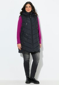 Žena v dlouhé černé zateplené vestě s kapucí, fialovém topu s dlouhým rukávem, vybledlých černých džínách a černých kotníkových botách، stojící na bílém pozadí.