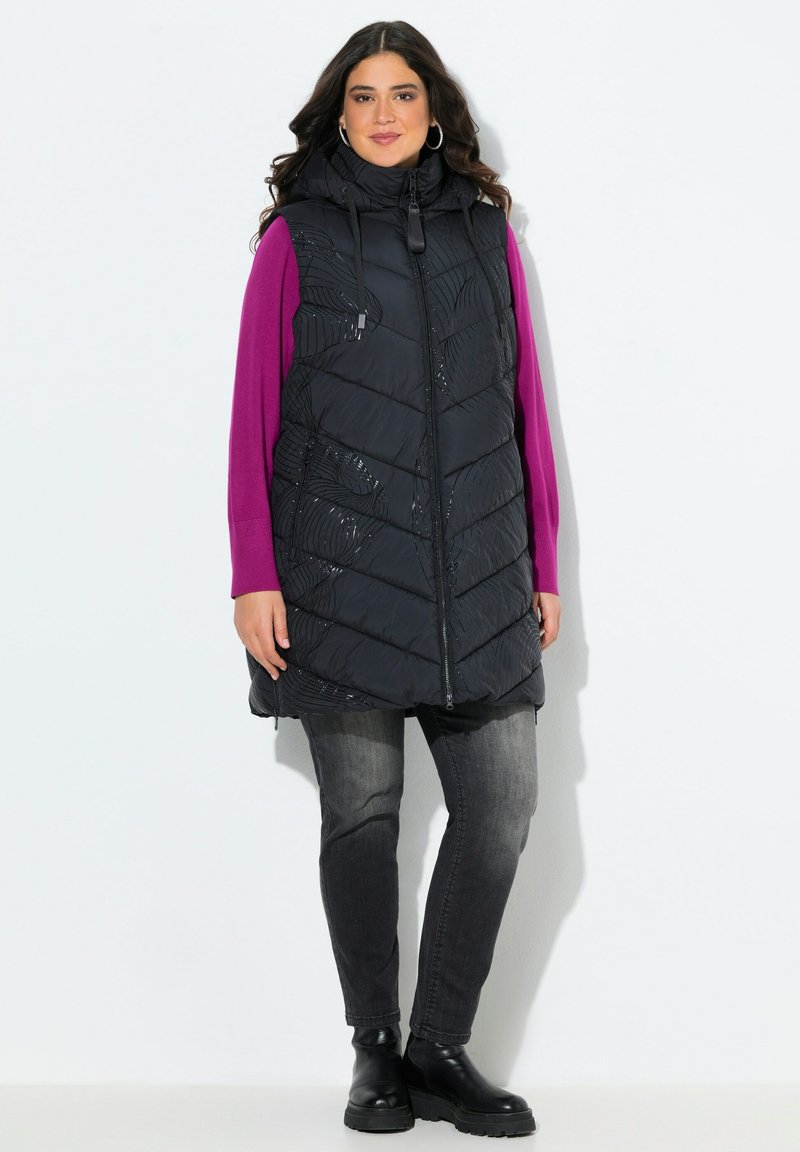 Žena v dlouhé černé zateplené vestě s kapucí, fialovém topu s dlouhým rukávem, vybledlých černých džínách a černých kotníkových botách، stojící na bílém pozadí.