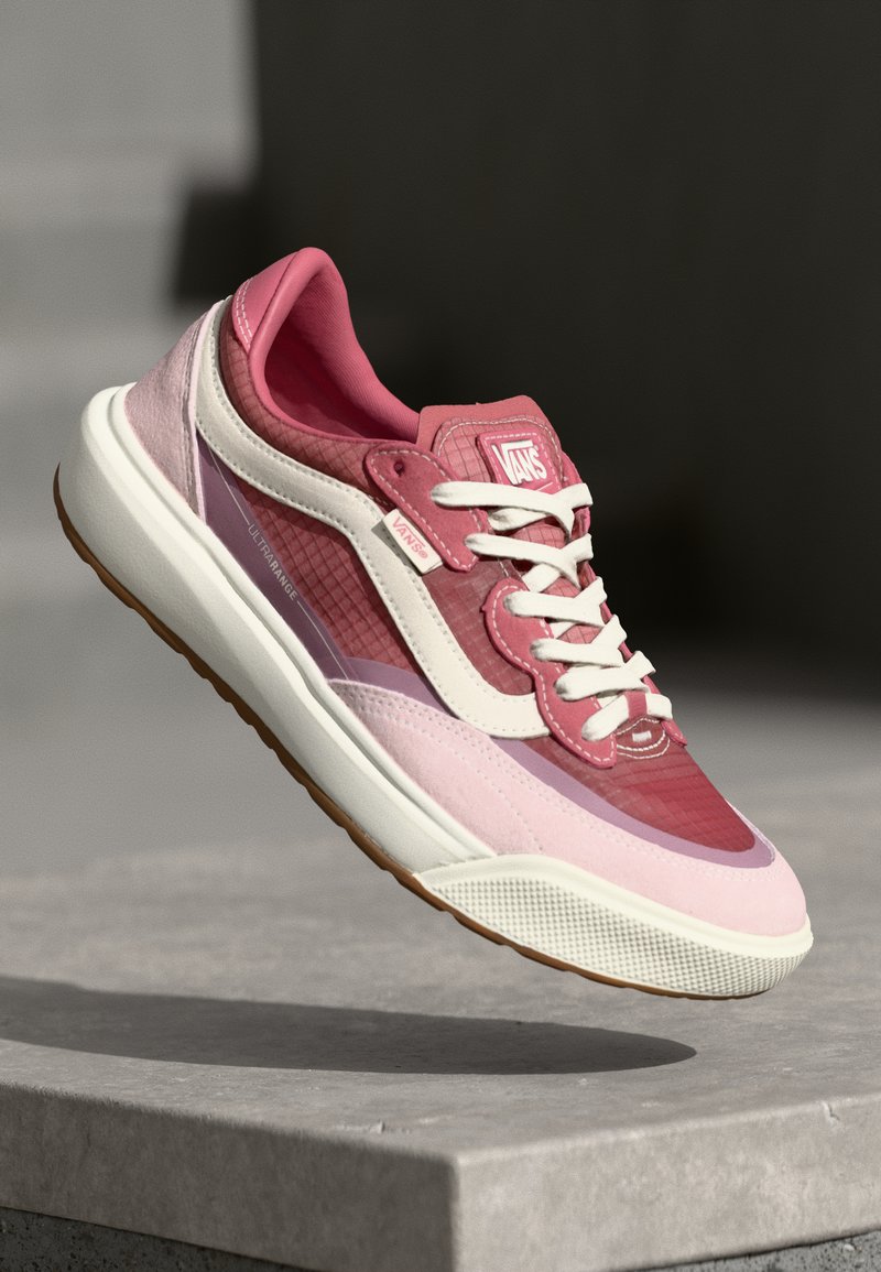 Adidași roz și roz deschis cu texturi din plasă și piele întoarsă, șireturi albe, talpă din cauciuc și elemente de design stratificate. Prezintă logo-ul Vans.