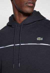Sweatshirt preto com capuz e riscas brancas, apresentando "LACOSTE" e um logótipo de crocodilo verde. Feito de um tecido suave com cordões.