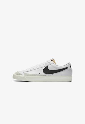 Valkoiset matalavartiset lenkkarit, joissa on musta Nike-logon swoosh, harmaa narskuva kärki ja valkoiset nauhat; kumipohja, jossa on teksturoitu yksityiskohta sivuilla.