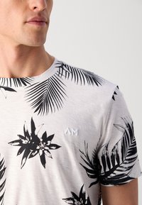 Witte T-shirt met zwarte tropische bladerenprint. Heeft een ronde halslijn en een klein geborduurd logo op de borst. Zachte, gladde textuur.