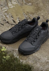 Scarpe da trekking nere con lacci appoggiate su un terreno fangoso con erba e una piccola pozzanghera vicino.