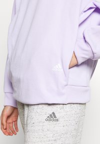Světle fialová mikina se žebrovanými manžetami a lemem, s malým bílým logem Adidas. Doplňují ji šedé strukturované tepláky s logem.