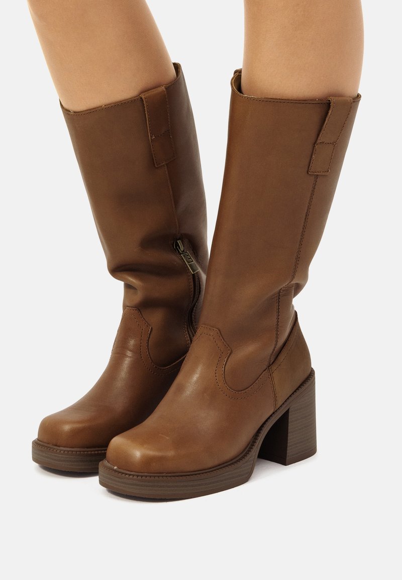 Bruine leren laarzen tot de knie met een chunky hak, ritsdetails en decoratieve stiksels. Het ontwerp heeft een ronde neus en een gladde textuur.