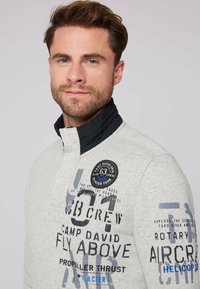 Maglione grigio con colletto alto, accenti tessuti in navy e grafiche testuali stampate. Presenta un distintivo tondo sulla parte sinistra del petto.