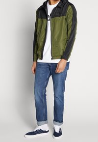 Olivengrøn og sort vindjakke med lynlås foran, justerbar hætte og elastiske ærmer. Båret over en hvid skjorte og blå jeans.