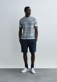 Man stands with hands in pockets wearing patterned short-sleeve shirt, dark shorts, dark socks, and white sneakers against plain background.