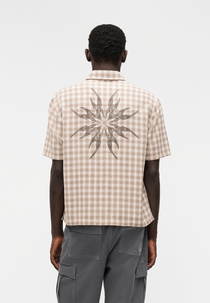 Πουκάμισο με κοντά μανίκια από μπεζ καρό ύφασμα, με μεγάλο γραφικό σχέδιο στην πλάτη. Μίνιμαλ γιακάς και ελαφρώς φαρδιά εφαρμογή.