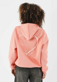 Sudadera color coral con un bolsillo frontal, capucha con cordón, y un diseño de corazón blanco en la parte posterior. La tela parece suave y texturizada.