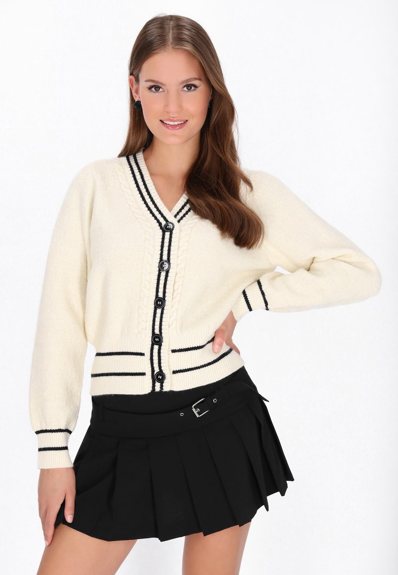 Cremefarbener Cardigan mit schwarzen Streifen, strukturiertem Strickmuster und sechs Knöpfen. Kombiniert mit einem schwarzen, plissierten Rock mit einem Gürtel.