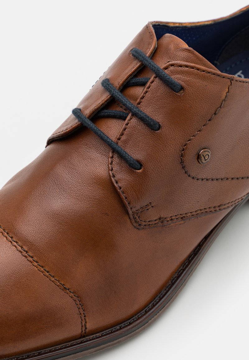 Zapato de cuero marrón con una textura lisa, que presenta cordones negros, una puntera cosida y un acento de logo metálico en el lateral.