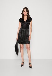 Robe en satin noir avec de courtes manches à capuche, décolleté en V et des boutons décoratifs sur le devant ; stylée avec des escarpins noirs à bout pointu et un sac.