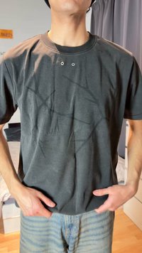 Muž v tmavosivej tričku s jemnými geometrickými vzormi a svetlomodrých džínsach stojaci v interiéri s rukami pri vreckách.