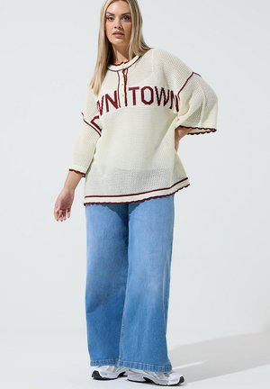 Kvinde stående iført en løs cremefarvet striktrøje med teksten "DOWNTOWN", vide blå jeans og hvide sneakers mod en ensfarvet baggrund.