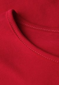 Detailaufnahme von rotem Stoff, die die feine Naht entlang einer gefalteten Kante zeigt und die Textur sowie die Nahtkonstruktion hervorhebt.