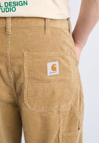 Primer plano de unos pantalones de pana color beige con un parche del logo de Carhartt en el bolsillo trasero, combinados con una camisa beige que presenta texto verde.