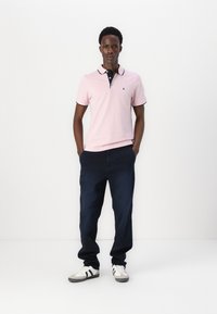 Roze polo shirt met marineblauwe kraag en rand, donkerblauwe broek en witte sneakers met zwarte strepen. Model staat met handen in de zakken.
