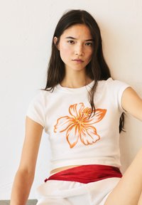 Witte t-shirt met korte mouwen en een groot oranje bloemenpatroon, gecombineerd met een rode stoffen riem. Glad textuur, getailleerde pasvorm.