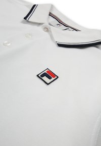 Witte polo met een marineblauw en rood logo op de borst en een marineblauwe kraag met knopen.