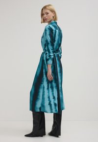 Abito di satin turchese con vita a nastro, maniche lunghe e una silhouette fluida. Caratterizzato da una combinazione di motivi blu chiaro e scuro.