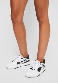 Hvite og svarte sneakers med gummisåle, med polstret krage, perforert tåboks og fremtredende svarte detaljer og logo.