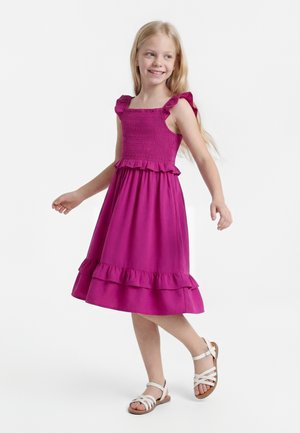 Mladé dievča sa usmieva a kráča, má na sebe ružové šaty bez rukávov s volánmi a biele sandále so remienkami na bielom pozadí.