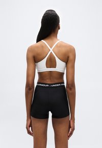 Γυναίκα που φοράει λευκό αθλητικό σουτιέν με σταυρωτό σχέδιο και μαύρο σορτς Under Armour, στέκεται γυρισμένη με την πλάτη προς τα έξω μπροστά σε απλό φωτεινό φόντο.