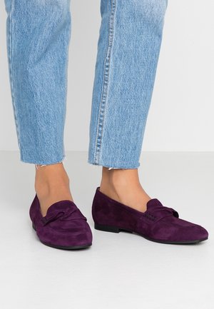 Voeten dragen paarse suède loafers gecombineerd met lichtblauwe cropped denim jeans op een witte vloer en achtergrond.