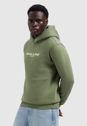 Man die een olijfgroene hoodie draagt met de tekst "BALLIN'", lichtblauwe spijkerbroek, staand tegen een effen lichtgrijze achtergrond.