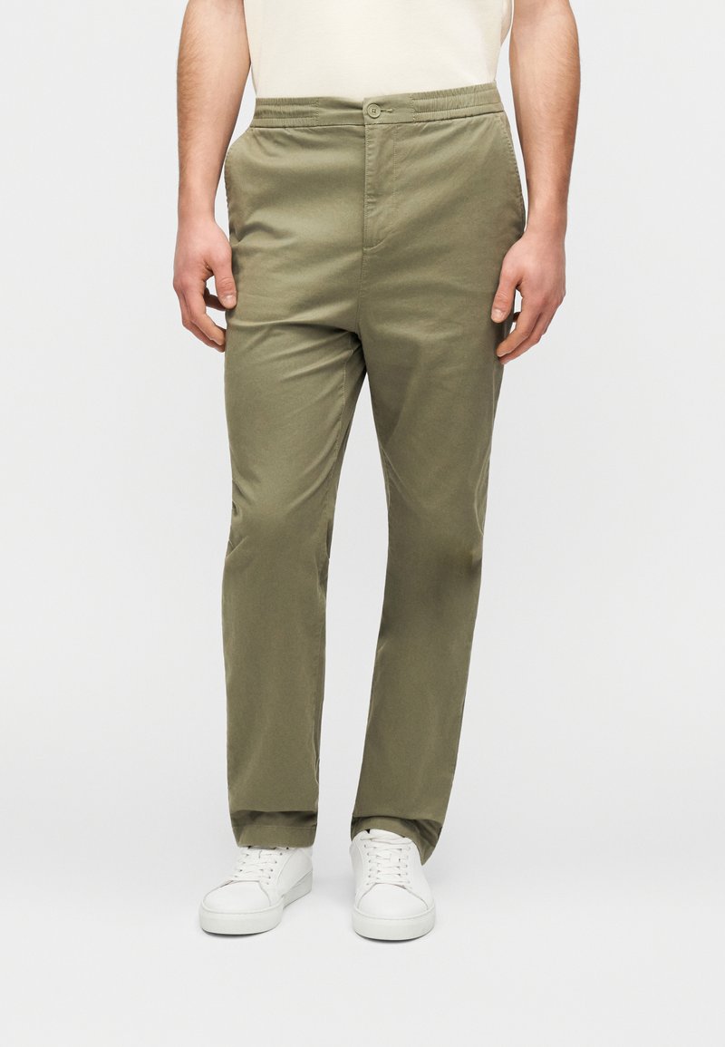 Pantalon vert olive à taille élastique avec fermeture boutonnée devant, doté d'une coupe droite et d'un tissu coton lisse. Associé à des baskets blanches.