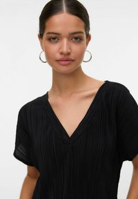 Giovane donna con i capelli scuri raccolti, che indossa orecchini a cerchio argentati e una maglia nera a V con pieghe, su uno sfondo bianco semplice.