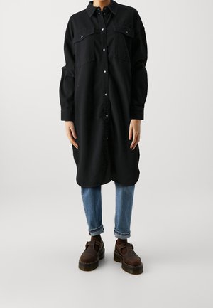 Person, die ein langes schwarzes Hemdkleid mit Knöpfen, hellblaue hochgekrempelte Jeans und dunkelbraune klobige Schuhe vor einem einfarbigen hellen Hintergrund trägt.
